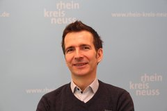 Das Bild einen Mann mit kurzen, dunkelbraunen Haaren. Er hat blaue Augen und ein freundliches, offenes Lächeln. Sein Gesichtsausdruck wirkt positiv und selbstbewusst. Der Mann trägt einen dunkelgrauen V-Ausschnitt-Pullover über einem Hemd. Der Hintergrund des Bildes ist ein hellblaues, einfarbiges Banner mit dem Logo "Rhein-Kreis Neuss" in weißer Schrift.