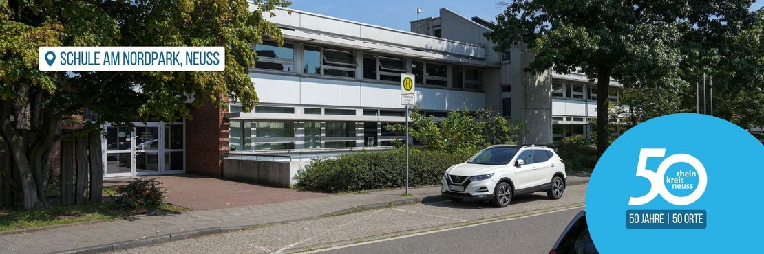 Älteres Schulgebäude, das von großen Bäumen eingefasst ist. Vor dem Schuleingang befindet sich eine Bushaltestelle für den Schulbus. Es parken Autos an der Straße vor der Schule.