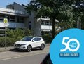 Älteres Schulgebäude, das von großen Bäumen eingefasst ist. Vor dem Schuleingang befindet sich eine Bushaltestelle für den Schulbus. Es parken Autos an der Straße vor der Schule.