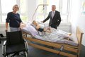 Landrat Hans-Jürgen Petrauschke steht an einem Krankenbett indem eine Pflegepuppe leigt. Frau Groth steht vor dem Bett, hinter einem leeren Rollstuhl