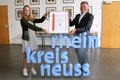 Madita Beeckmann und Landrat Hans-Jürgen Petrauschke stehen hinter einem hüfthohen Rhein-Kreis Neuss Logo und halten die Urkunde mit dem Gütezeichen hoch.