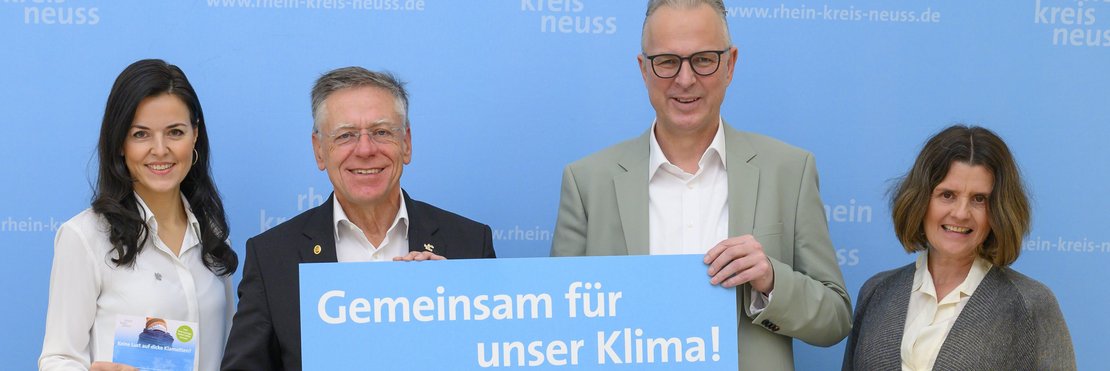 Landrat Hans-Jürgen Petrauschke (2.v.l.) mit Kreisdirektor Dirk Brügge und den Klimaschutzmanagerinnen Ina Grothe (l.) und Regina Krings vor einer blauen Wand mit Rhein-Kreis Neuss Logo. Die beiden Herren in der Mitte halten ein Schild mit der Aufschrift "Gemeinsam für unser Klima" hoch.