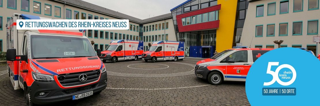 Das Bild zeigt 8 Krankenwagen, die vor einer Rettungswache aufgestellt sind. Davor stehen quer zwei Notarzt-Fahrzeuge. In der Mitte der Fahrzeuge befindet sich eine Gruppe von Mitarbeitenden der Rettungswache.