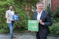 Landrat Hans-Jürgen Petrauschke und Thiago de Carvalho Zakrzewski stehen lächelnd, mit Abstand zueinander, vor einer grünen Hecke und halten Schilder in Ihren Händen. Auf diesen steht: "Gesundheit und Wohlergehen" und "Partnerschaften zur Erreichung von Zielen"