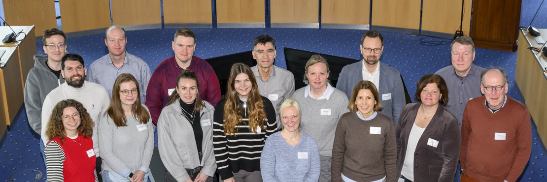 Das Bild zeigt eine Gruppe von 15 Personen, die in einem Sitzungssaal für ein Gruppenfoto posieren. Die Personen sind unterschiedlich alt und tragen zwanglose Kleidung. Sie sind unterschiedlich groß und haben verschiedene Frisuren und Haarfarben. Viele lächeln in die Kamera. Die meisten tragen Namensschilder.