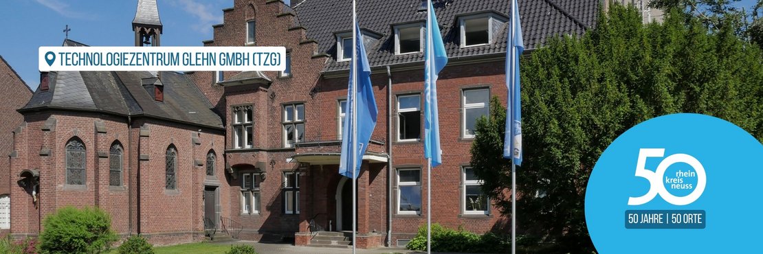 Historisches Backsteingebäude, im Hintergrund strahlend blauer Himmel. Vor dem Gebäude stehen drei blaue Flaggen, die das Logo des Rhein-Kreis Neuss zeigen.