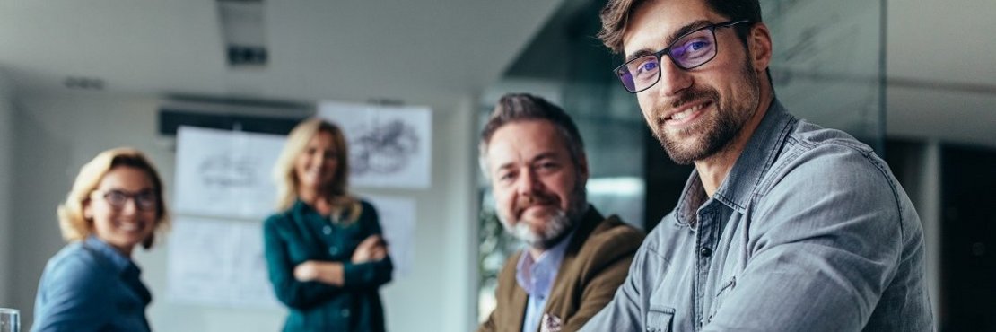 Vier Personen in einem Meeting an einem Tisch. Sie schauen in die Kamera.