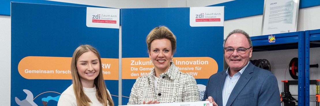 Zwei Frauen und ein Mann vor einer blauen Wand mit einem Schild in der Hand.