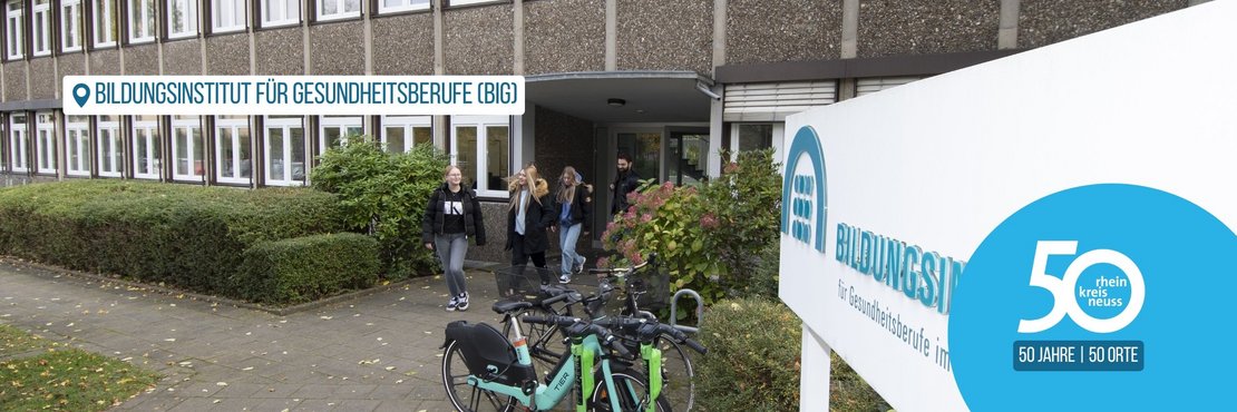 Das Bild zeigt vier junge Leute, die das Bildungsinstitut für Gesundheitsberufe (BIG) in Neuss verlassen, zwei E-Bikes stehen davor,  und ein Schild mit der Aufschrift Bildungsinstitut für Gesundheitsberufe im Rhein-Kreis Neuss. Das Bild zeigt eine moderne, graue Betonfassade eines Gebäudes mit vielen großen Fenstern. Vor dem Gebäude befindet sich ein gepflegter, grüner Strauchheckenstreifen. Vier junge Erwachsene, zwei Frauen und zwei Männer, verlassen das Gebäude. Sie sind lässig gekleidet; zwei der Frauen tragen Jacken. Vor ihnen stehen zwei helltürkise E-Bikes, die an einem Fahrradständer angelehnt sind. Ein großes, weißes Schild steht rechts im Bild, auf dem in blauer Schrift „Bildungsinstitut für Gesundheitsberufe im Rhein-Kreis Neuss“ steht. Ein großer, hellblauer Kreis enthält die Zahl „50“ und die Worte „50 Jahre | 50 Orte“.  Die Gesamtatmosphäre ist ruhig und alltäglich, die Stimmung neutral bis leicht positiv, da die jungen Menschen das Gebäude verlassen und der Jubiläumstext auf dem Schild positive Assoziationen weckt.