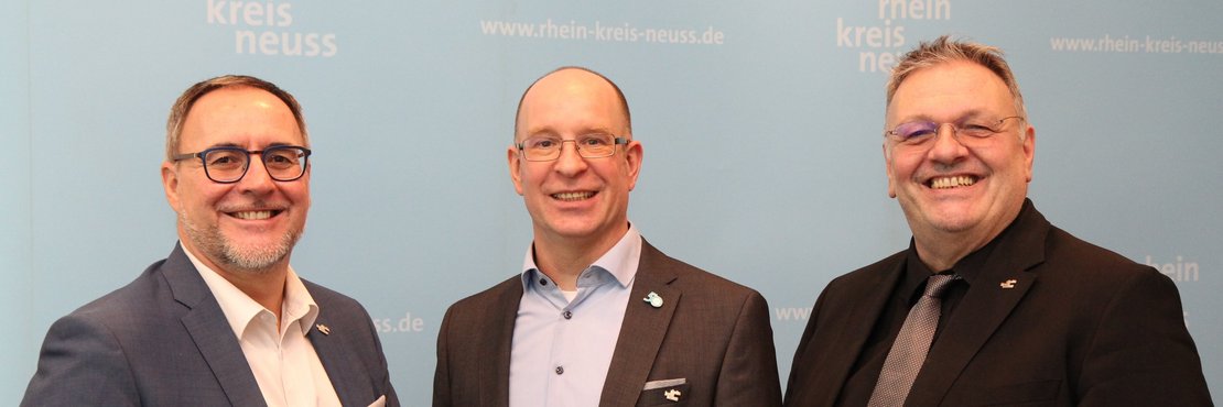 Das Bild zeigt drei Männer, die vor einer hellblauen Wand stehen. Die Wand trägt das Logo des Rhein-Kreises Neuss sowie die dazugehörige Webseite. Der Mann ganz links trägt einen dunkelblauen Anzug mit hellem Hemd und hält ein Tablet in der Hand. Er trägt eine Brille und hat kurz geschnittenes, braunes Haar. Der Mann in der Mitte trägt einen dunkelgrauen Anzug mit hellblauem Hemd und hat ebenfalls eine Brille. Der Mann rechts trägt einen schwarzen Anzug mit hellem Hemd und einer dunkelgrauen Krawatte. Er trägt ebenfalls eine Brille und hat kurz geschnittenes, graues Haar. Alle drei Männer lächeln in die Kamera.