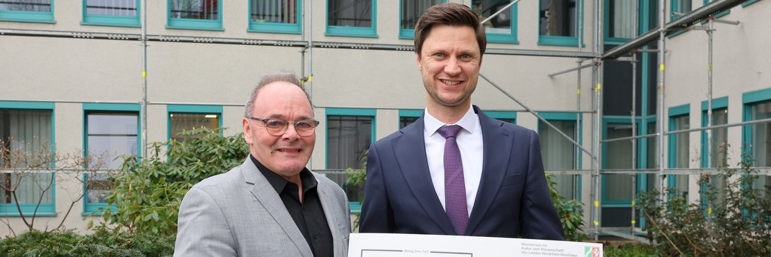 Das Bild zeigt zwei Männer, die vor einem Gebäude mit Gerüst stehen. Der Mann links trägt ein hellgraues Sakko über einem dunklen Hemd und eine Brille. Er hält mehrere Broschüren in der Hand, auf denen "MINT" steht. Der Mann rechts trägt einen dunkelblauen Anzug mit einem lilafarbenen Hemd und einer Krawatte. Er hält einen großen Scheck, auf dem "210.000 Euro" zu lesen ist, zusammen mit dem Empfänger "Netzwerk Rhein-Kreis Neuss".