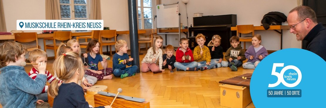 Ein Musiklehrer unterrichtet eine Gruppe von Vorschulkindern auf dem Boden in einem hellen Klassenzimmer.  Das Bild zeigt eine Gruppe von etwa zwölf Kindern im Vorschulalter, die in einem Kreis auf dem Holzfußboden sitzen. Die Kinder haben verschiedene Musikinstrumente wie Xylophone, Triangel und Trommeln und benutzen diese. Ein erwachsener Mann, ihr Lehrer, sitzt auf dem Fußboden und spielt ein Xylophon. Er trägt eine dunkle Strickjacke und Jeans und unterstützt die Kinder beim Musizieren. Die Atmosphäre im Raum ist entspannt und freundlich. Die Kinder scheinen interessiert und engagiert zu sein. Das Licht im Raum ist hell und natürlich, und die Gesamtstimmung ist warm und positiv. 